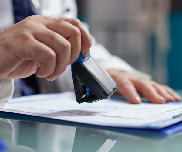 Doctor holding medical stamp to put on prescription paper at health care exam. Hand of physician putting seal on checkup report, giving medicine to patient at consultation. Close up